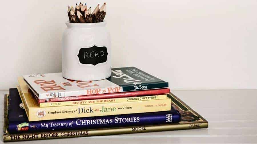 A stack of children’s books with a white vase filled with colored pencils on top.