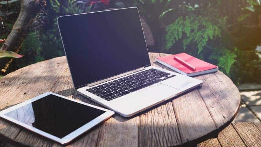 A wooden picnic table with a laptop, tablet,  notebook, and pen. 