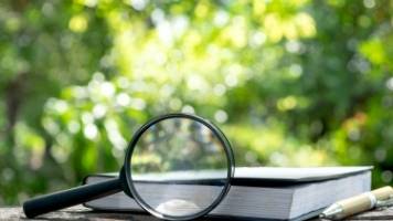 A book and a writing pen on top of a bench outside. A magnifying glass is propped up against the book. 