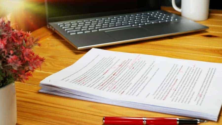 A laptop, coffee mug, and a stack of papers that have been proofread sit on a wooden desk. 