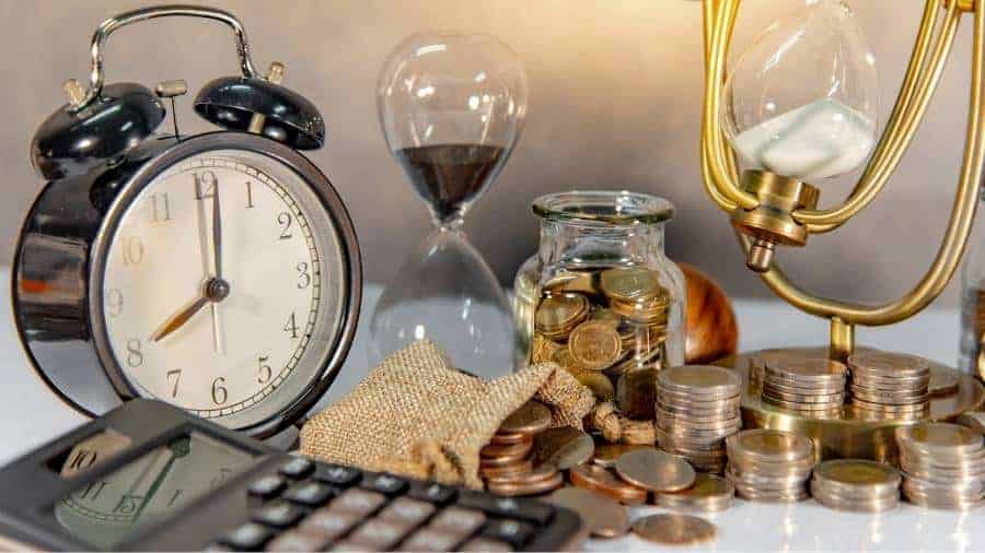 An alarm clock, calculator, sand timer, and stacks of coins sit on a desk. 