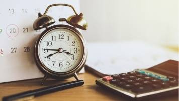 A calculator, an alarm clock, a calendar, and a black pen on top of a wooden desk. 