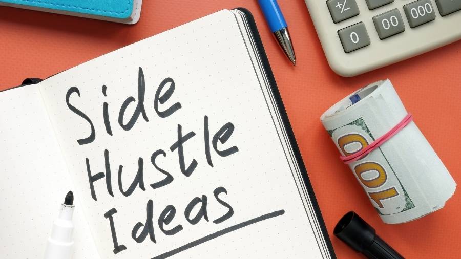 A calculator, a pen, a wad of money, and a notebook with the words "Side Hustle Ideas" written in black marker. 