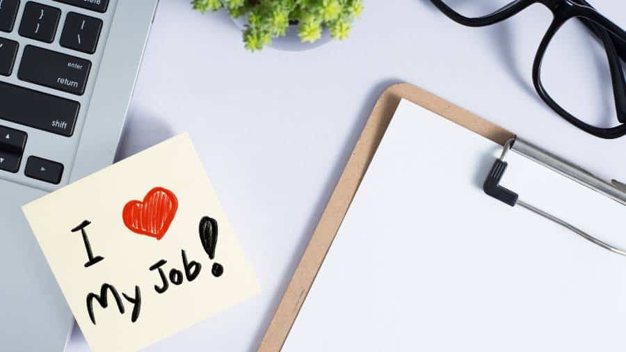 A white desk with a laptop and assorted items. A sticky note attached to the laptop says, "I Love My Job!" 