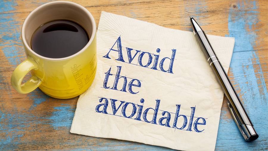 A coffee mug, writing pen, and napkin with the message "Avoid the avoidable" are sitting on a wooden table. 